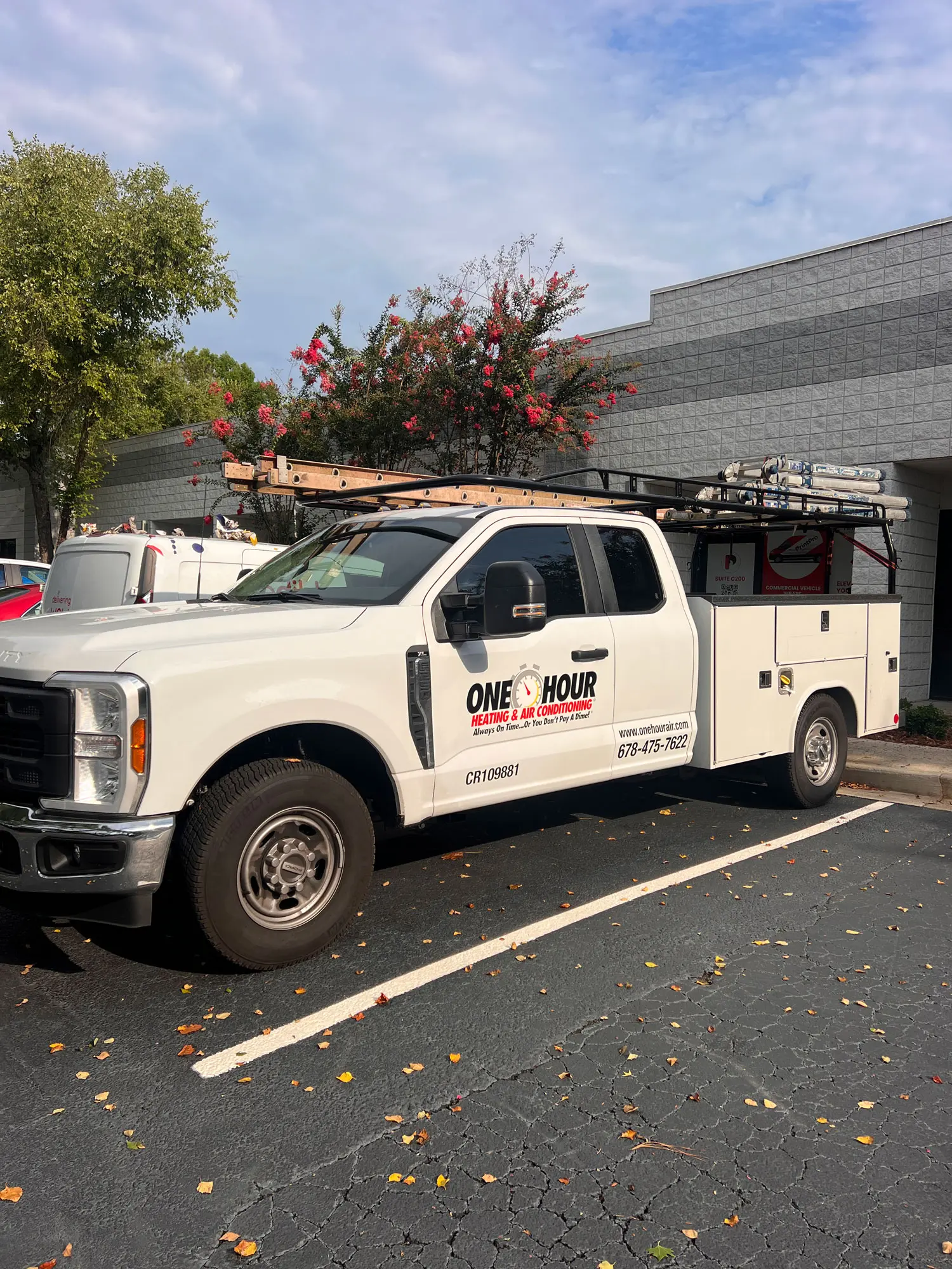 A white service truck with ladders on top, labeled "One Hour Heating & Air Conditioning," is parked in a lot near a gray building.