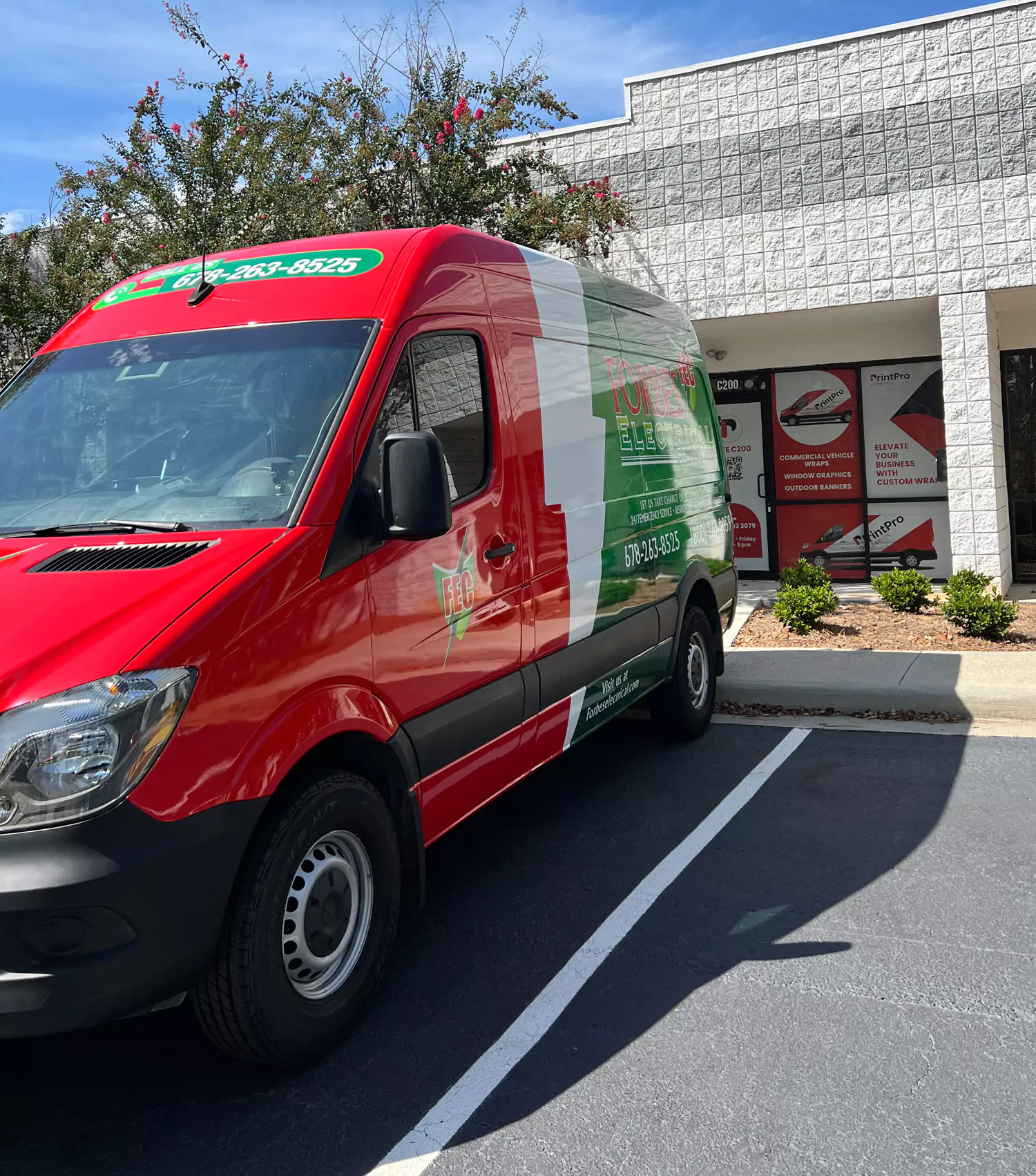 Red and white delivery van parked outside a brick building, with green signage on both the van and building. Bright, sunny day with clear skies.