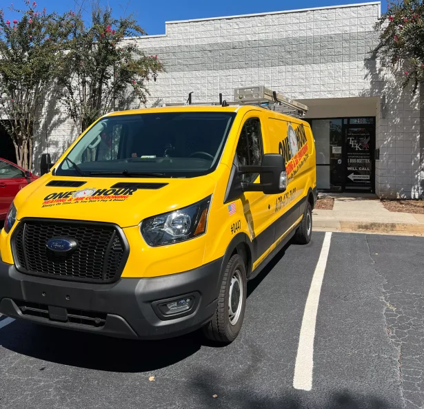 Bright yellow service van parked outside a white brick building. The van displays "One Hour" branding. Sunny day with shadows and greenery nearby.